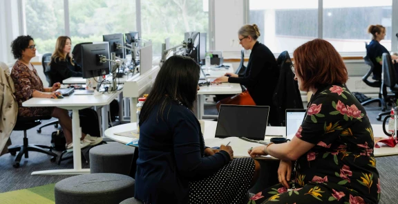 Two people sitting at a table with laptops in front of them, with people at desks in the background